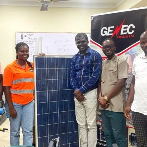Five people are standing indoors around a large solar panel, with a "GENEC Electrix Ltd." banner behind them. The setting is an office with technical drawings on a whiteboard in the background.