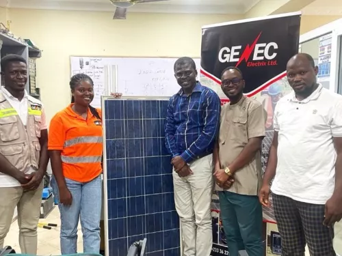Five people are standing indoors around a large solar panel, with a "GENEC Electrix Ltd." banner behind them. The setting is an office with technical drawings on a whiteboard in the background.